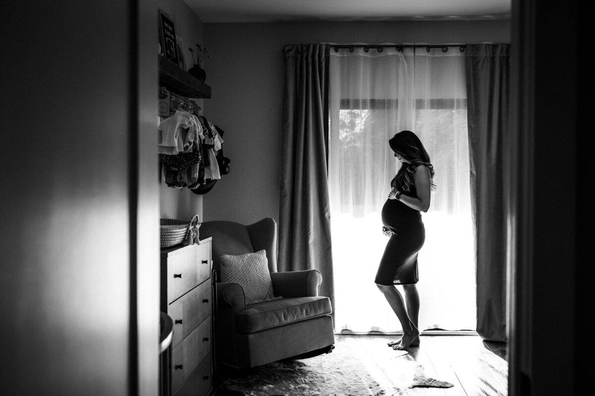 pregnant woman standing in nursery in front window with curtains looking down at her baby bump