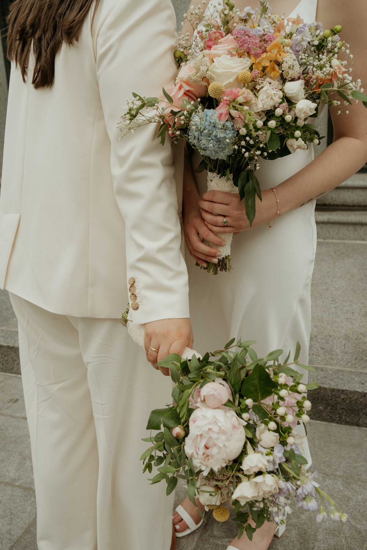 Close-up photo of a wedding couple holding bouquets. Both individuals are dressed in white; one wears a suit while the other wears a dress. The bouquets are filled with a variety of flowers, including peonies, roses, and hydrangeas, showcasing soft pastel colors. The image captures the elegant simplicity of their attire and the beauty of the floral arrangements against a stone staircase backdrop.