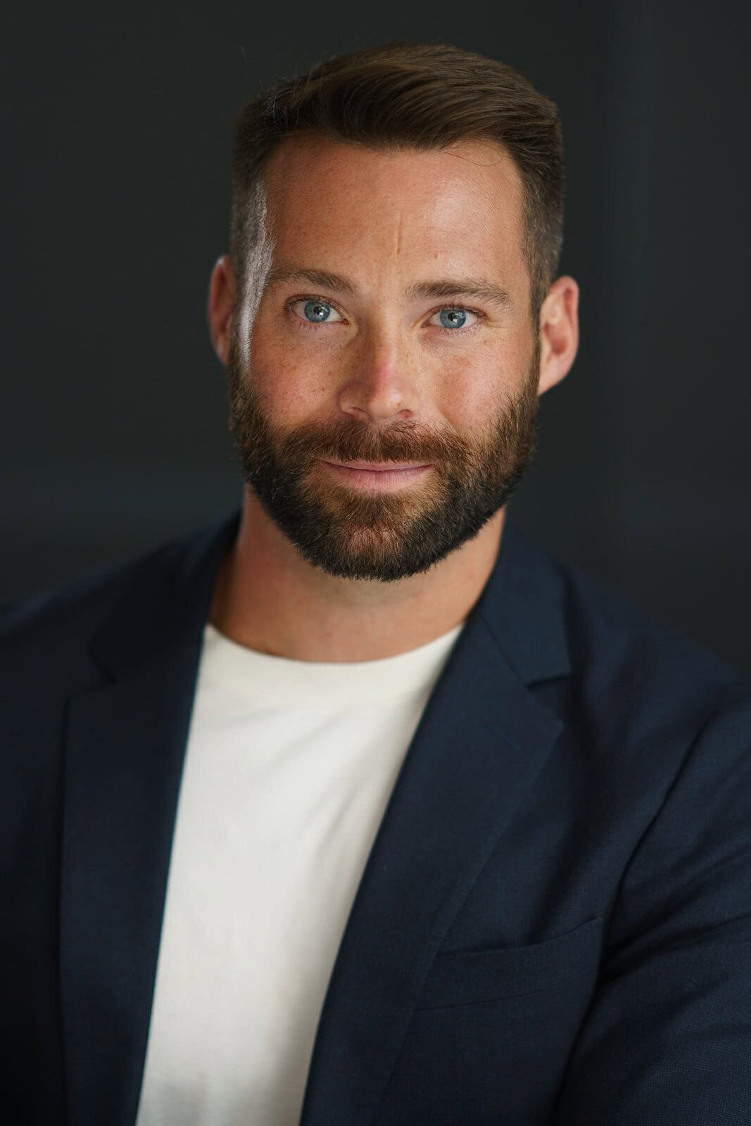 Young bearded man posing against a dark background while wearing a dark jacket and a white t-shirt