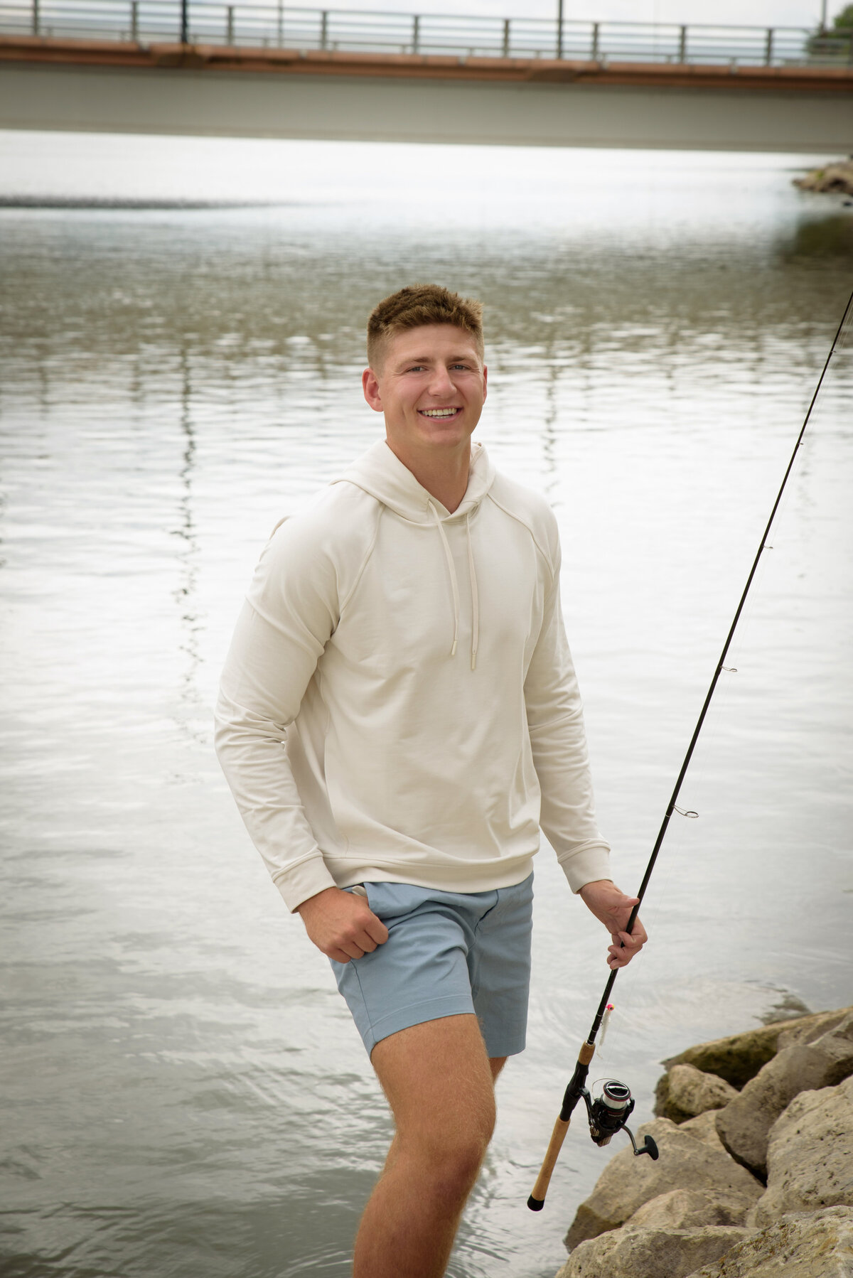 De Pere High School senior boy wearing blue shorts and a cream hooded sweatshirt sitting on rocks along the Fox River holding a fishing pole The Depot in Downtown Green Bay, Wisconsin