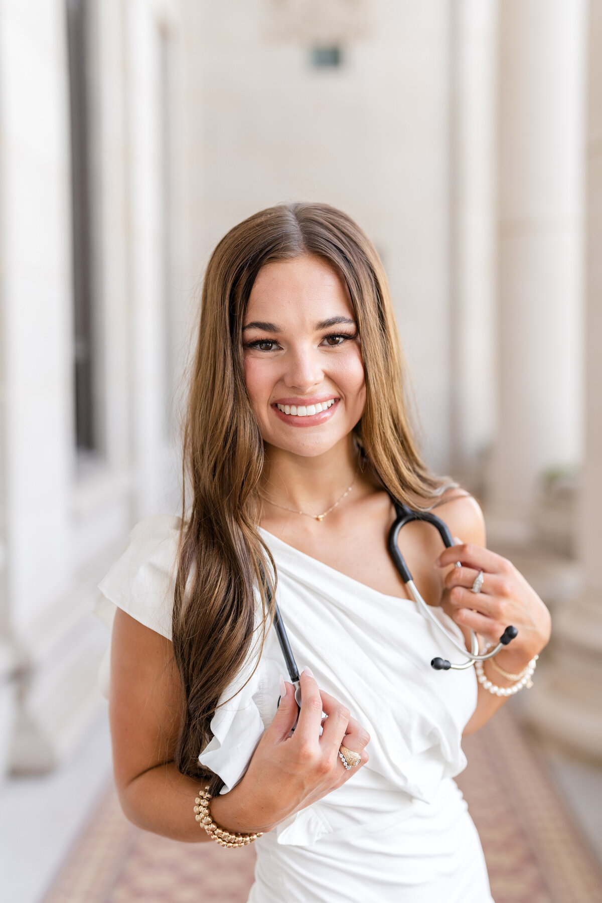 Texas A&M senior girl wearing stethoscope and white one shoulder dress while smiling in the columns of the Administration Building