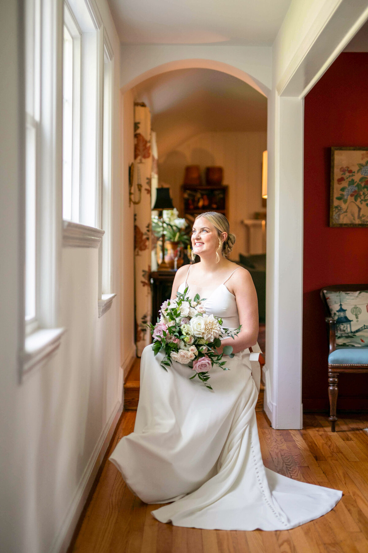 A bride in a white dress sits on a wooden floor near a window, holding a bouquet of pink and white flowers. She is smiling and looking out the window. A chair with a cushion is in the background, and the room features warm lighting.