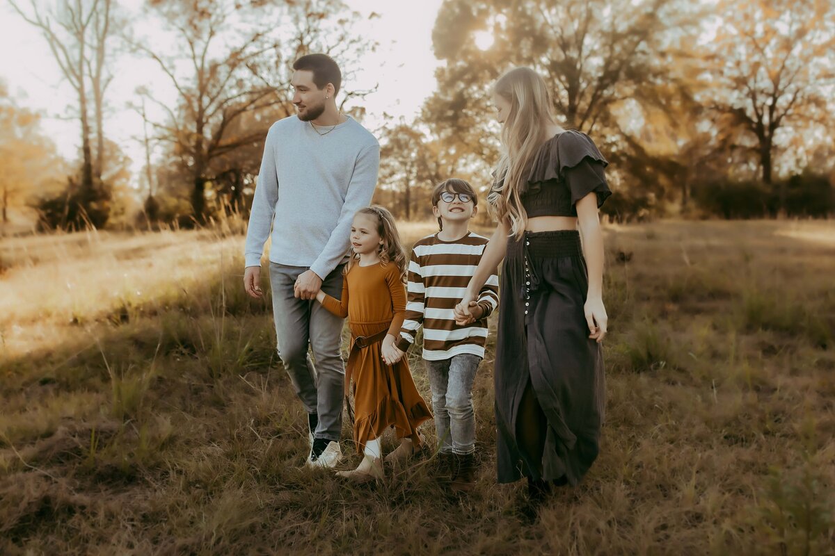 A family of four, two adults and two children, walks together in a grassy field with autumn trees in the background.