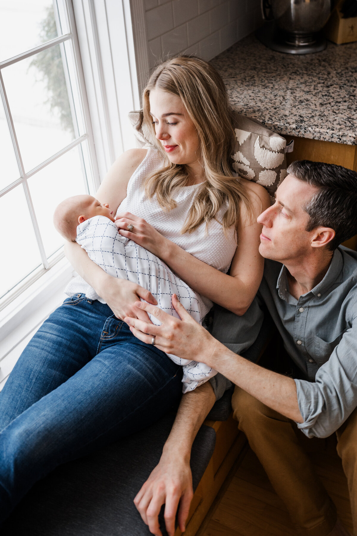 A mother rests on a window seat, cradling her swaddled newborn baby, while the father sits beside her, gently holding her arm.