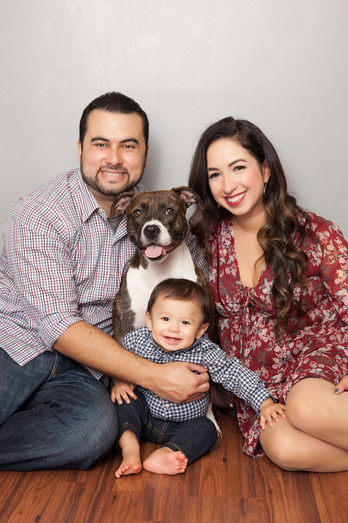 A man and woman, both smiling, sit on the floor holding a happy baby between them in this charming Jacksonville family photography session. Dressed casually, with the woman in a floral dress, they are joined by their cheerful brown and white dog sitting between them.