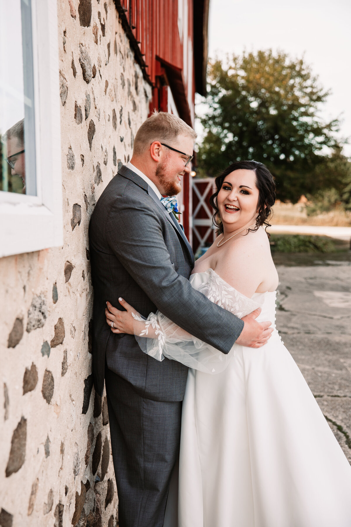 A bride laughs while hugging her groom against a stone wall