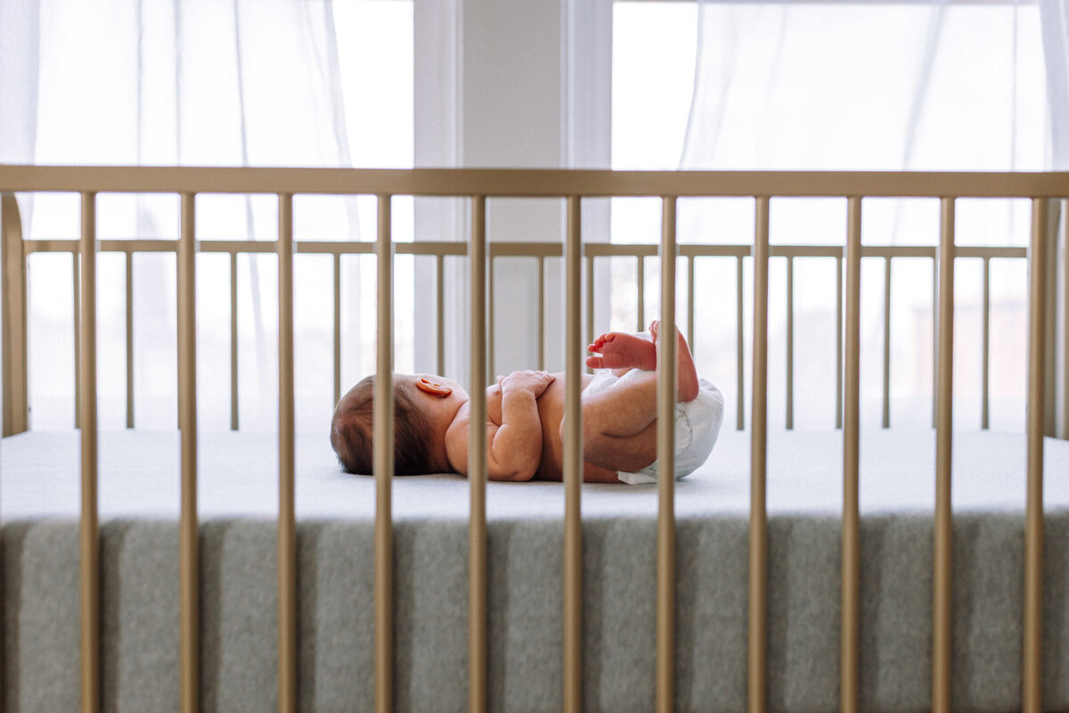 newborn in diaper resting in crib in front of two windows with curtains blowing in the wind