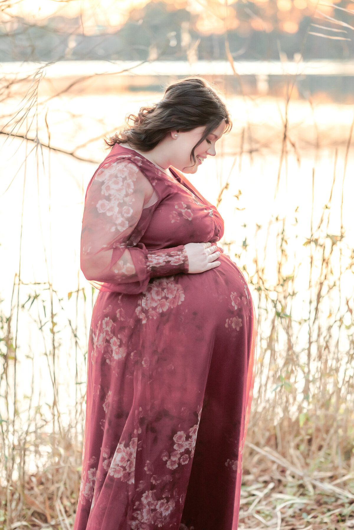 A mom-to-be looks down at her pregnant belly and smiles during her Chesapeake, VA maternity photography session. She wears a pink floral dress.