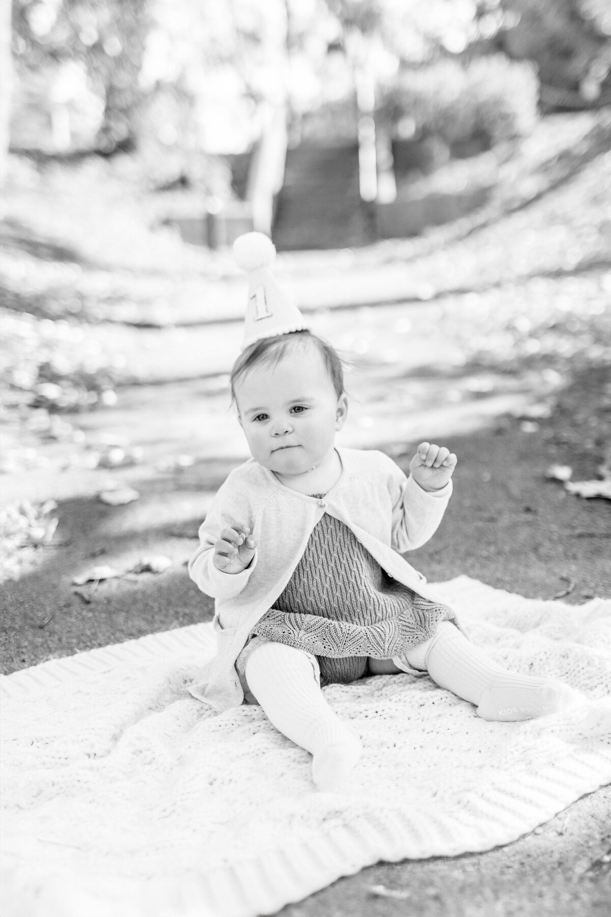 Black and white image of baby girl sitting on a knit blanket outdoors during her first birthday milestone session at a Kansas City park.