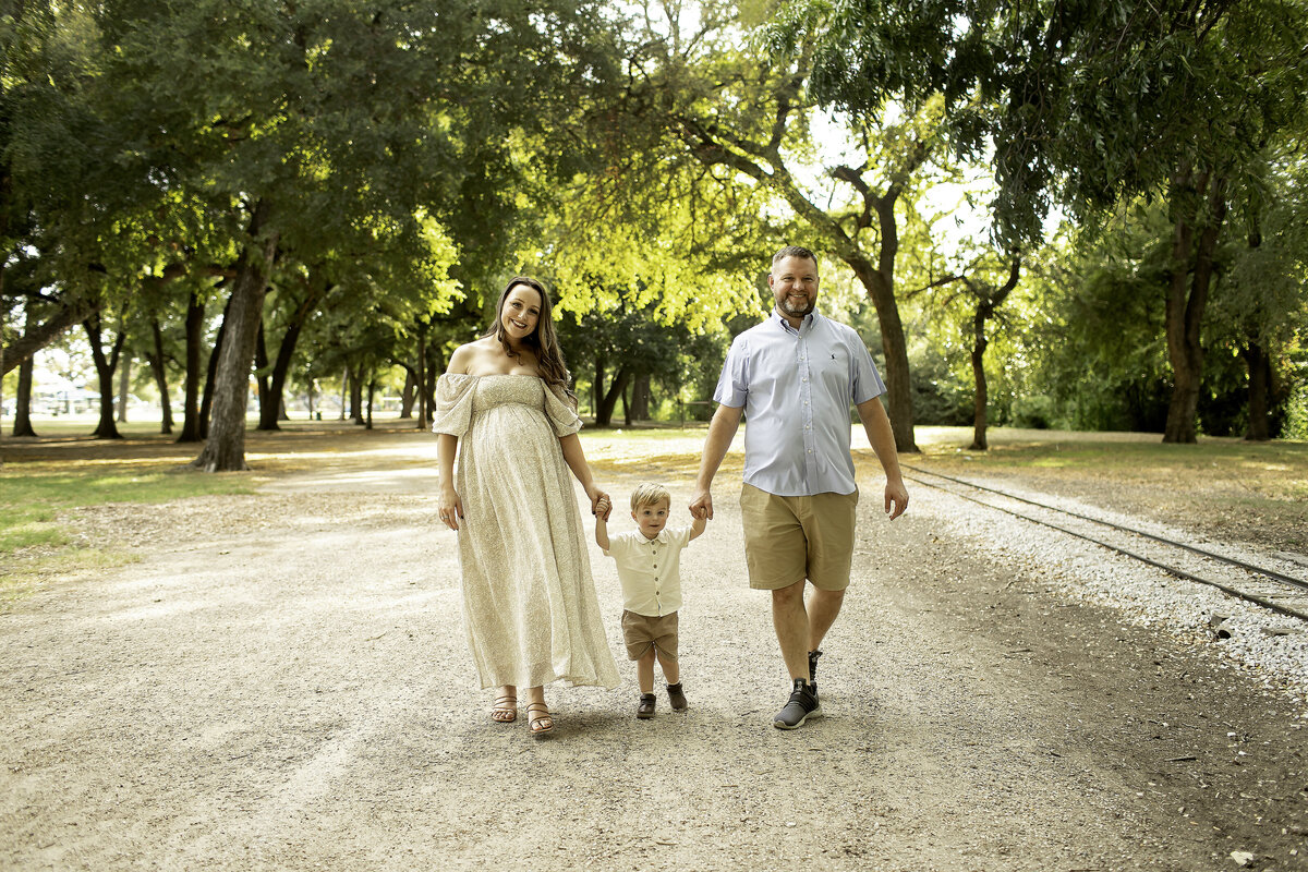 A joyful maternity photo at Trinity Park, with mom, dad, and big brother walking hand-in-hand along a scenic path, smiling and enjoying the moment as a family, captured by Chunky Monkey Photography.