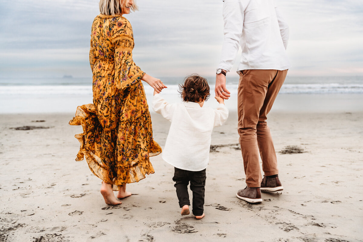 A man and woman hold hands with their toddler son and run toward the water, during a Coronado Beach photo session by lifestyle photographer Love Michelle Photography