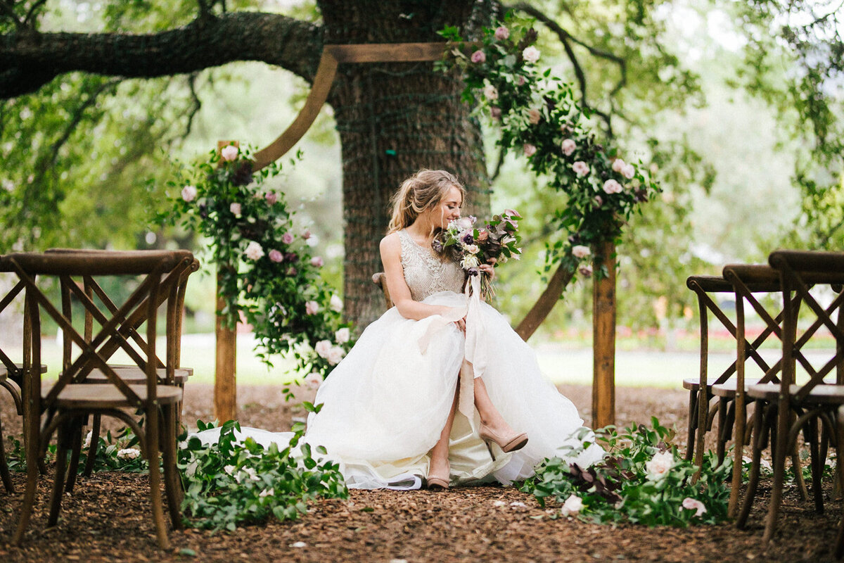 Bride sitting underneath a wooden wedding day ceremony arch decorated with florals and greenery
