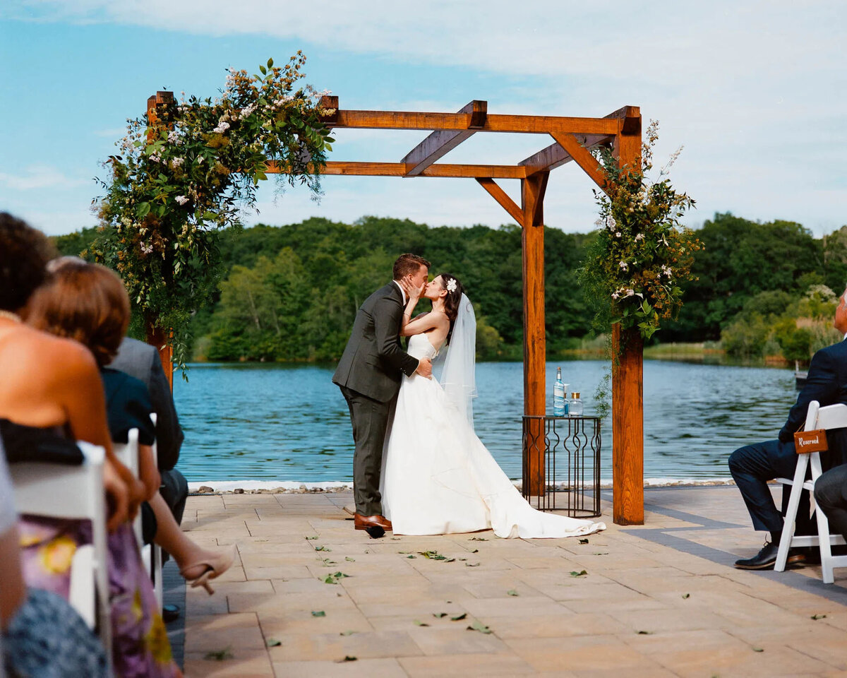 A bride and groom kiss under a wooden arch adorned with greenery and flowers by a lakeside, as guests watch their outdoor ceremony—captured beautifully by an NJ wedding photographer.