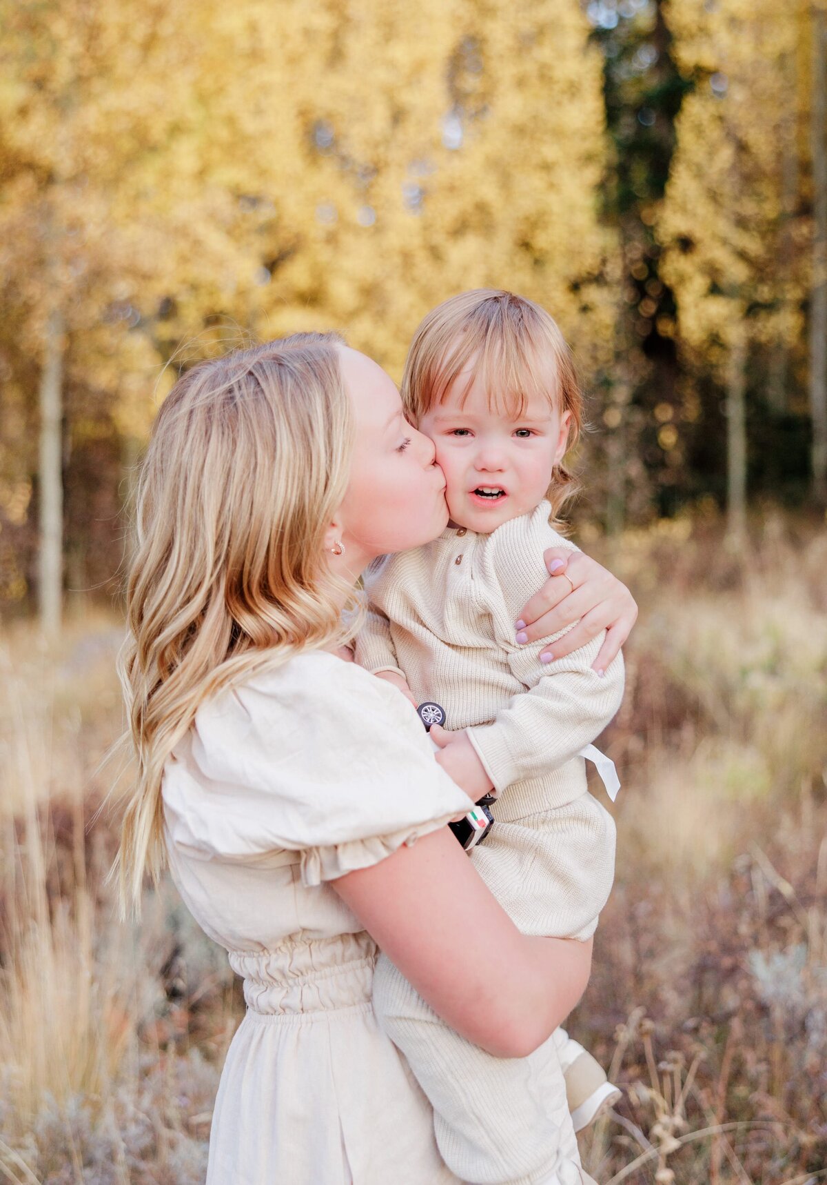 A teen girl in a cream dress kisses her baby sibling in a forest in fall