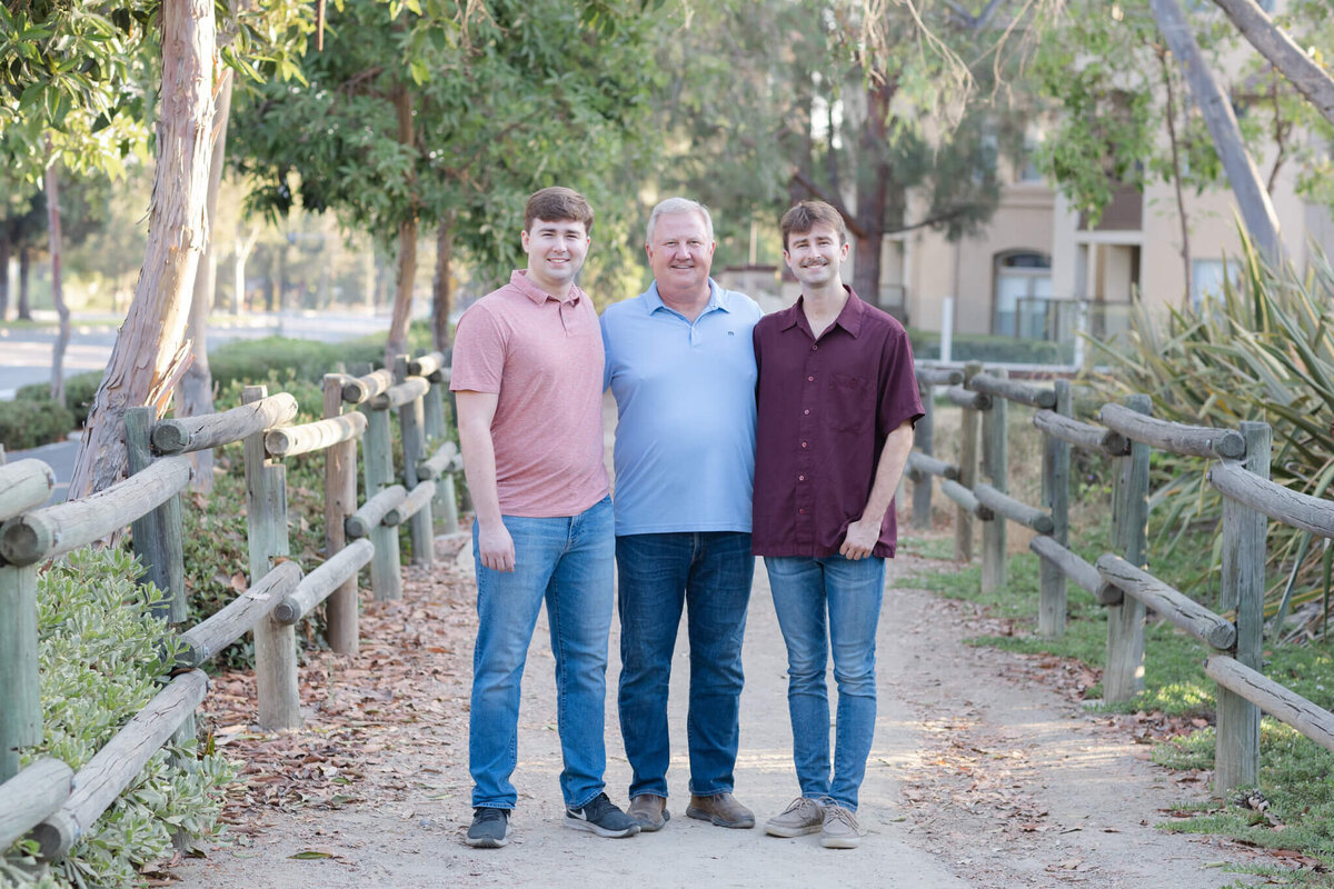 A father standing in the middle of his two adult sons with dirty blonde hair, putting his arms around them, standing in a gravel pathway surrounded by greenery.