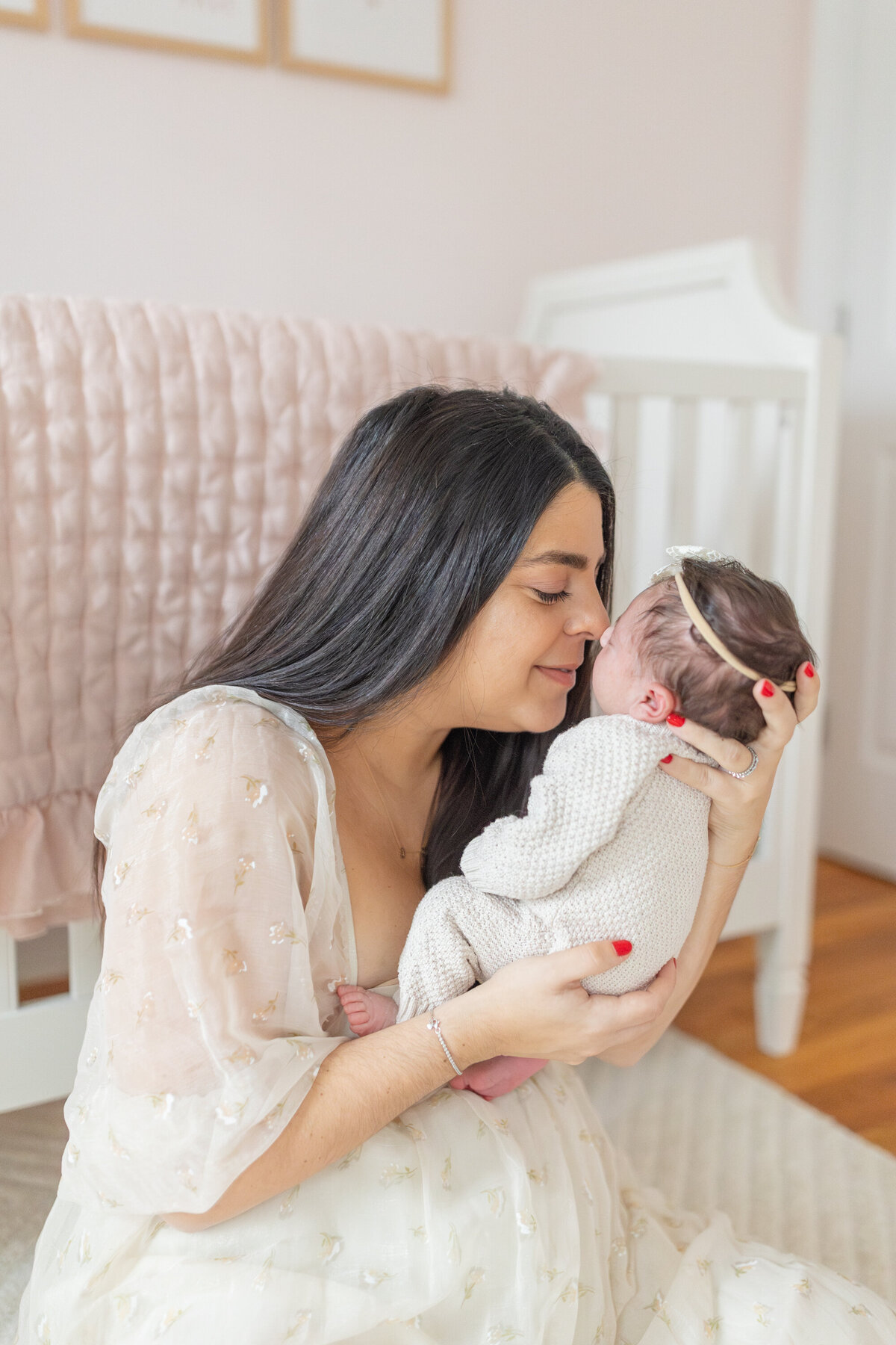 New mom sits on the floor in her baby's nursery, with the crib in the background, holding her newborn up to her face. She gently touches noses with her baby while smiling, capturing a tender and joyful moment of love and connection