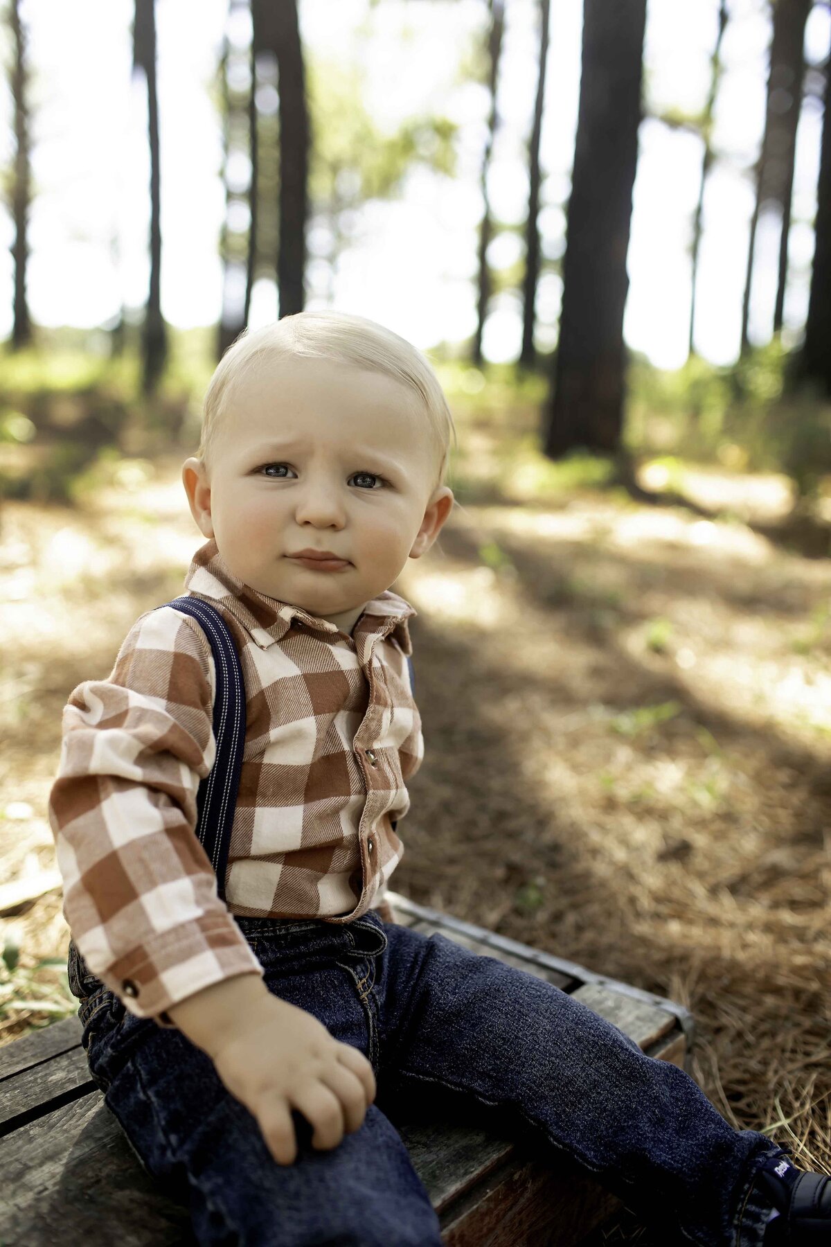 One-year-old birthday boy wearing overalls with suspenders, standing confidently in a pine tree forest