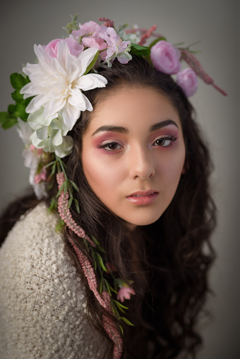 Editorial beauty portrait of a woman wearing soft glam makeup and a flower crown, posed elegantly in a studio with a clean backdrop.