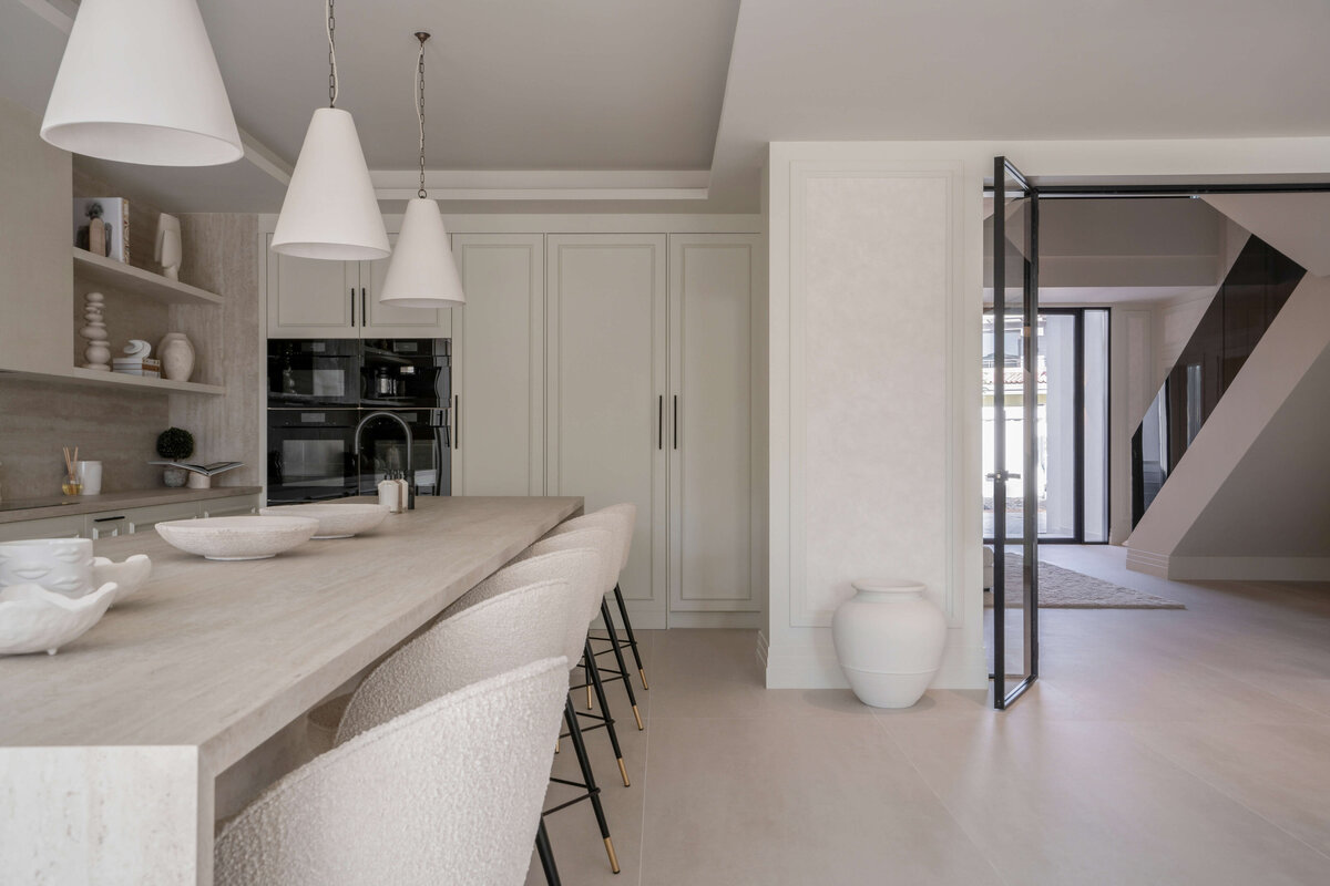 Spacious, open kitchen with  light, airy feel. To the left, a long  light colored bar with six white rounded barstools. Above, three white pendant lights. To the right, the dramatic front staircase is visible through open glass doors.