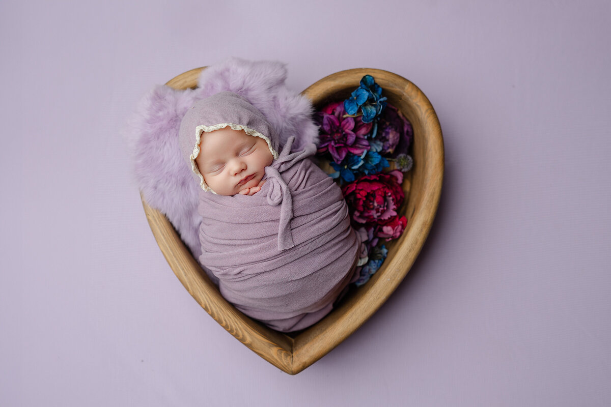 Baby girl styled in pastel purple tones with faux florals and fuzzy textures, resting peacefully inside a heart prop. Photographed by Inland Empire newborn photographer Kaitlyn Dawn.