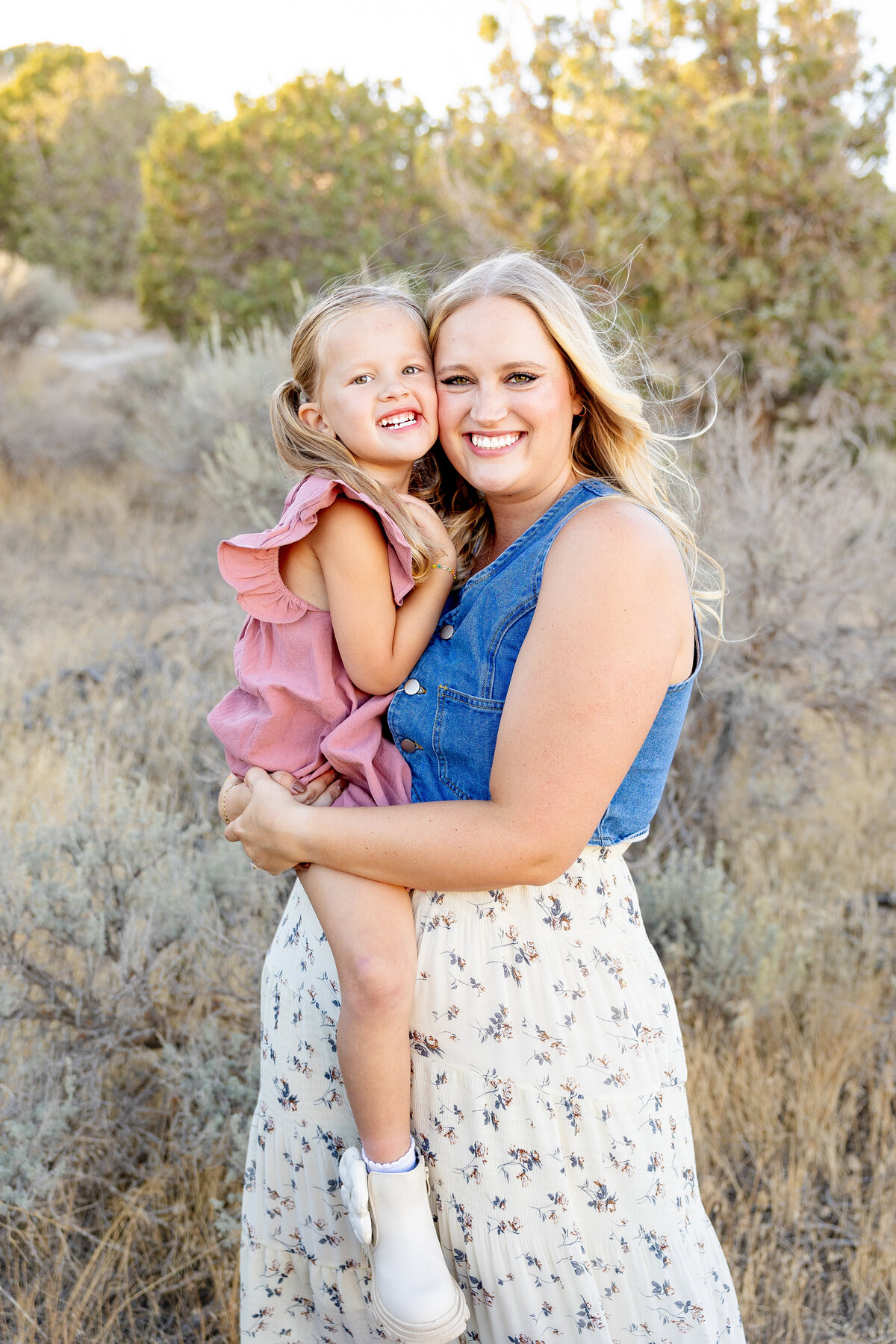 A mother in a blue denim top and floral skirt holds her young daughter, both smiling warmly against a backdrop of desert foliage and soft golden light.