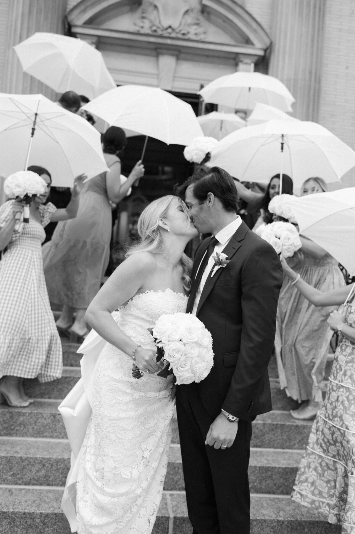 A bride and groom share a kiss on the steps of Spring Lake Golf Club, surrounded by friends and family holding white umbrellas and bouquets of white roses