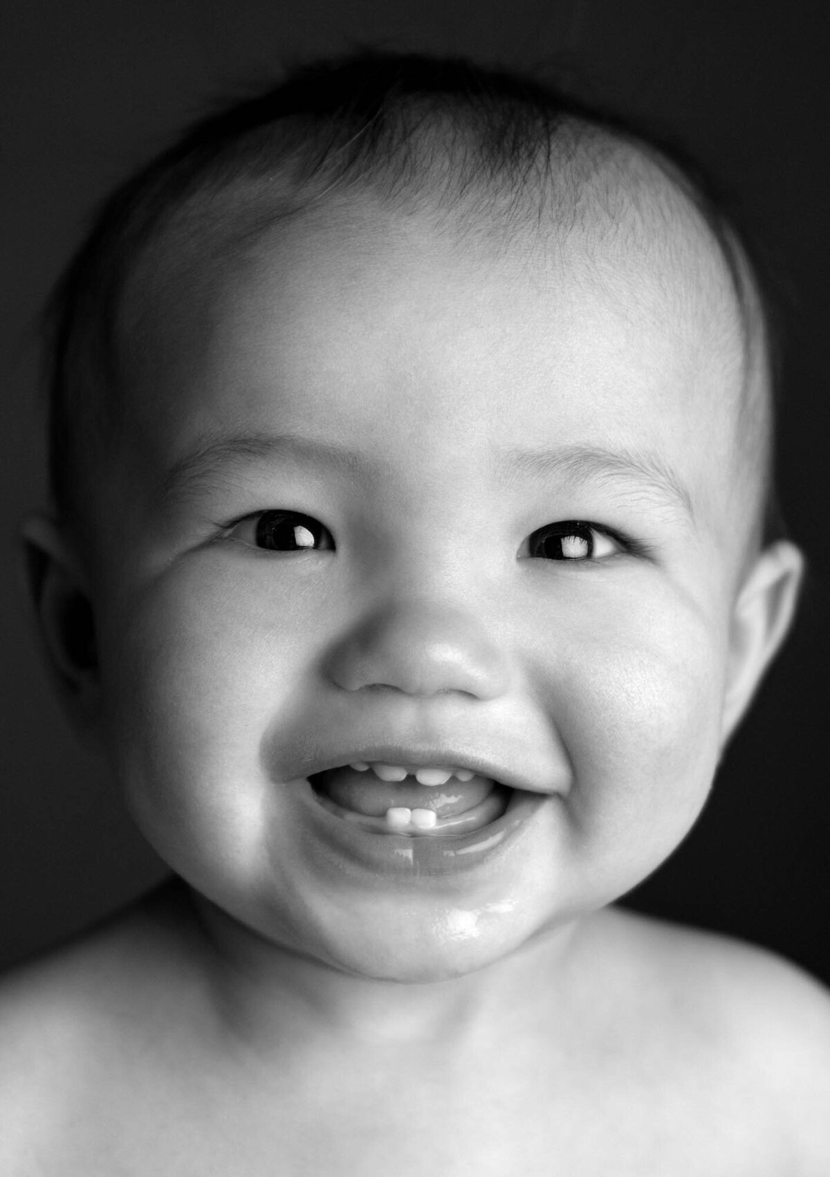 Black and white close-up of a joyful baby smiling widely, showing two tiny teeth. The baby's eyes sparkle with happiness against a dark background.