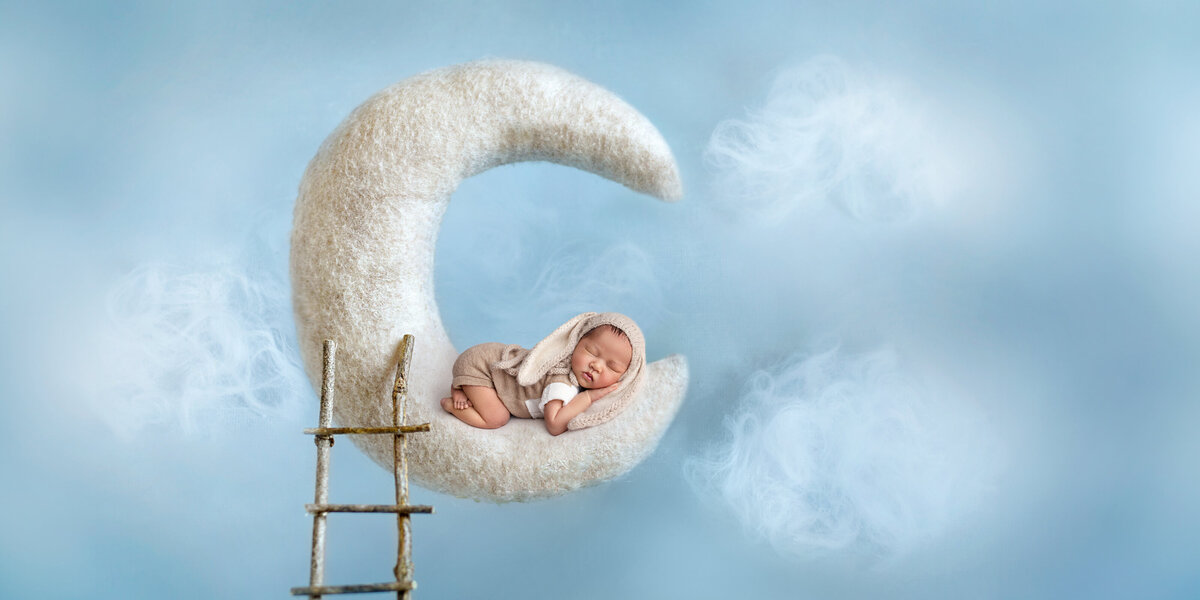 Sleeping newborn posed on a soft crescent moon prop with a tiny ladder, surrounded by fluffy cloud details on a blue sky background.