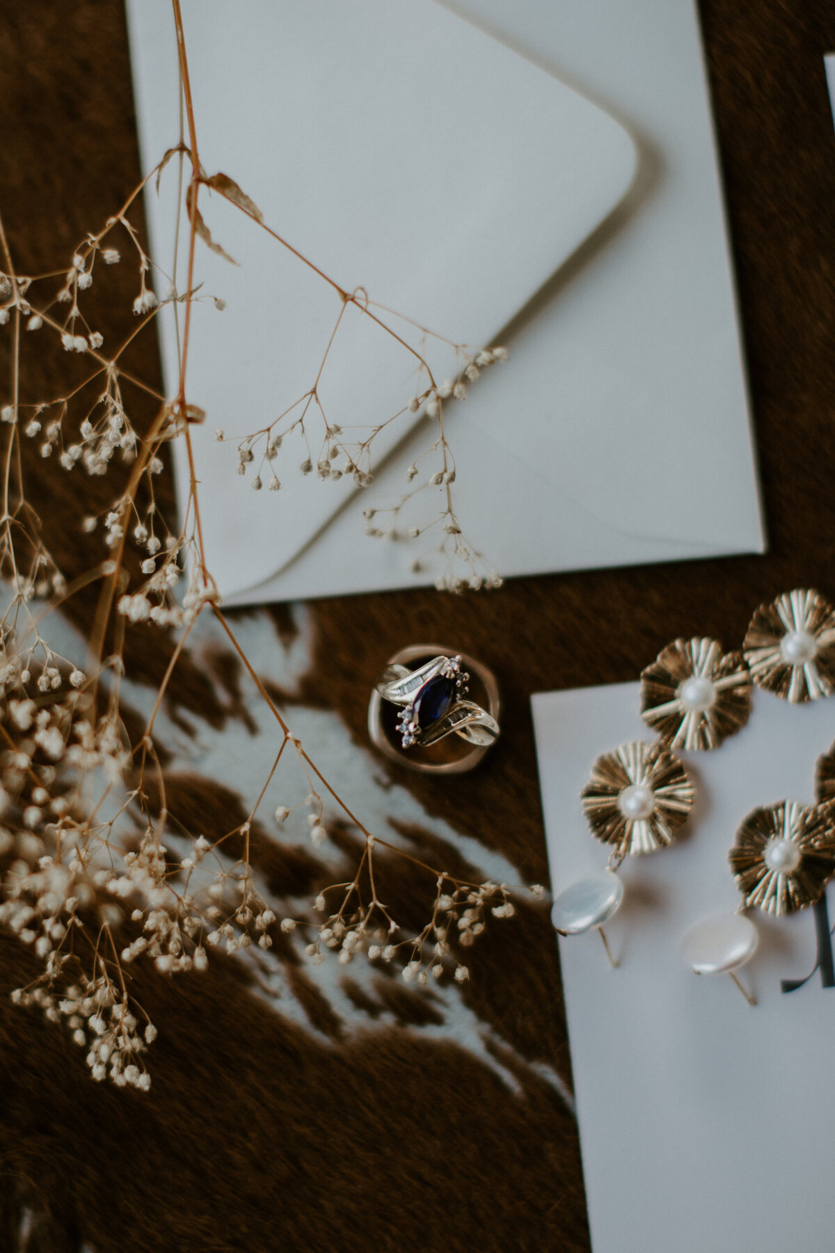 A pair of gold earrings and wedding bands on white stationery with dried flower stems atop cow hide.