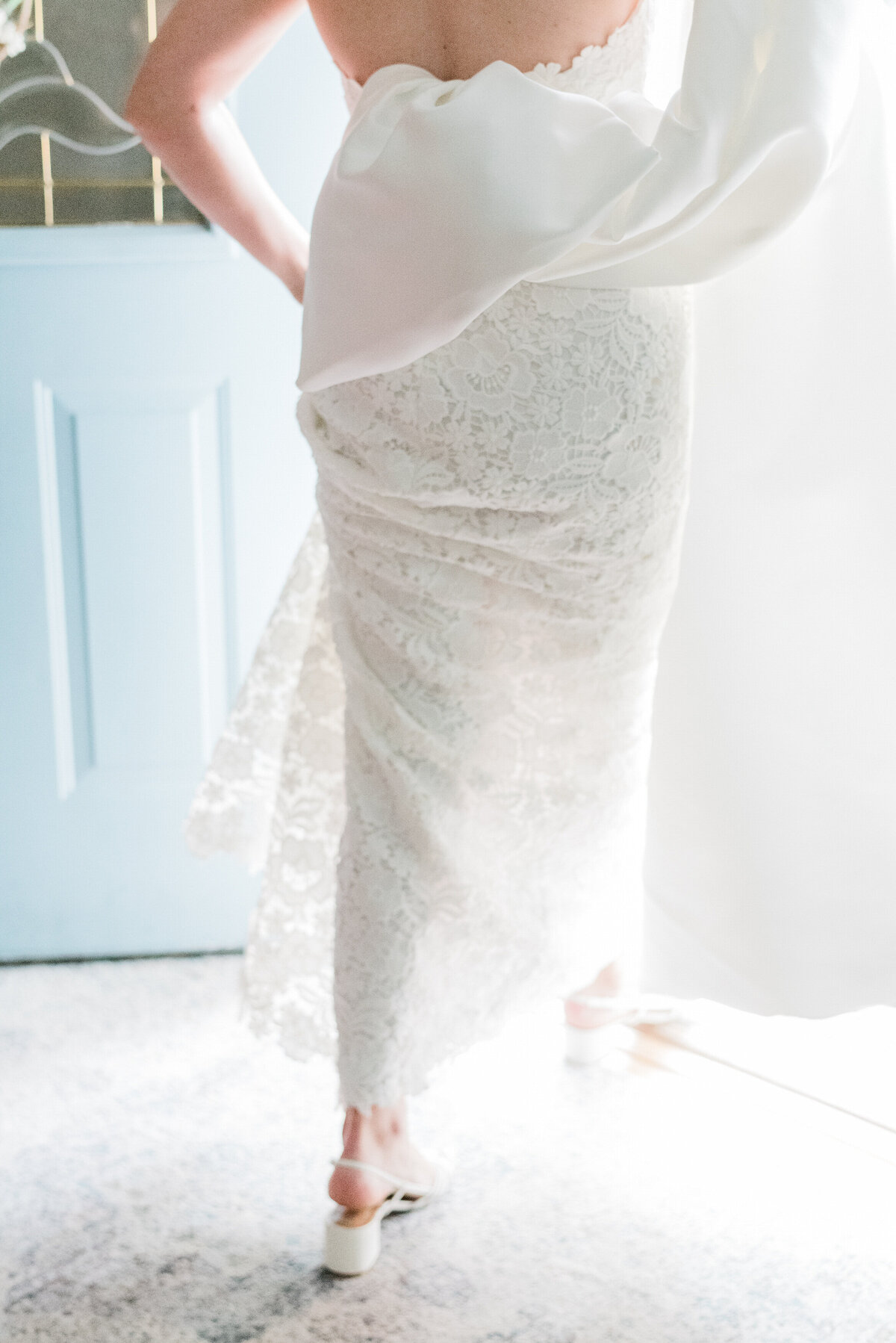 A close-up of the bride’s lace gown and statement bow as she steps through a doorway into a softly lit space.