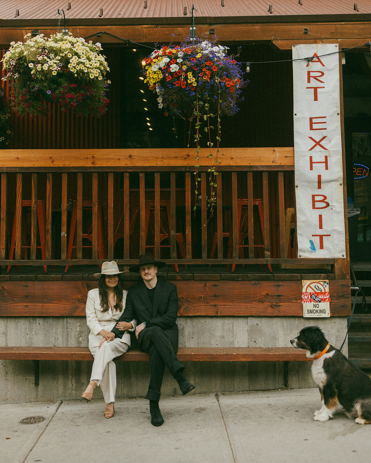 Couple sitting on bench with large flower baskets above them a dog is looking at them