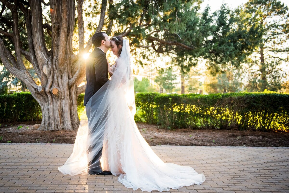 A groom kisses his bride on the forehead with the sun setting in the background.