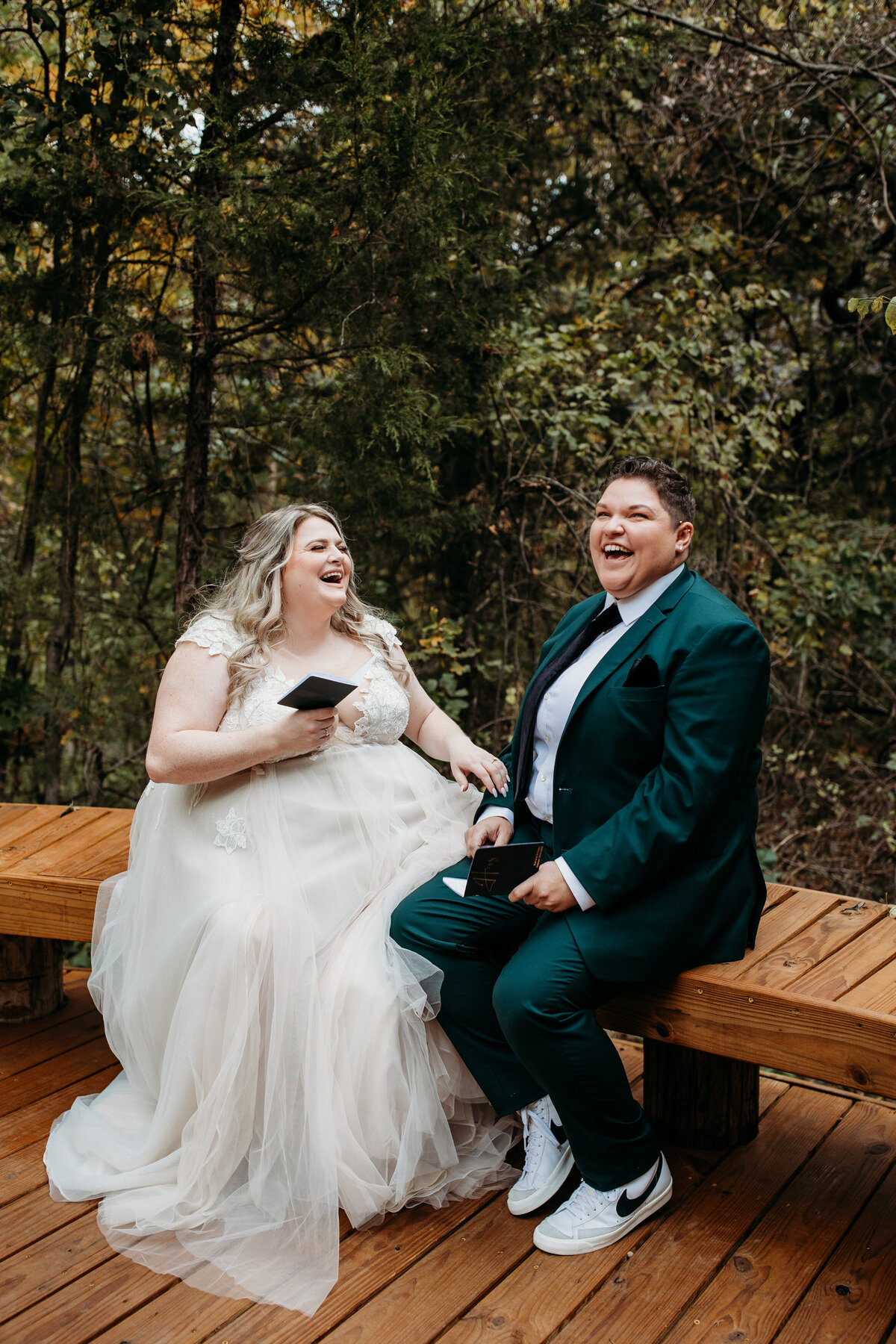 Two brides sharing a laugh while sitting on a wooden bench in a forested area