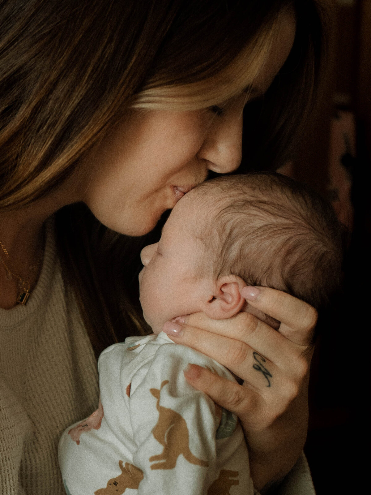 Mom gently kisses baby girl's forehead near soft window light at home.