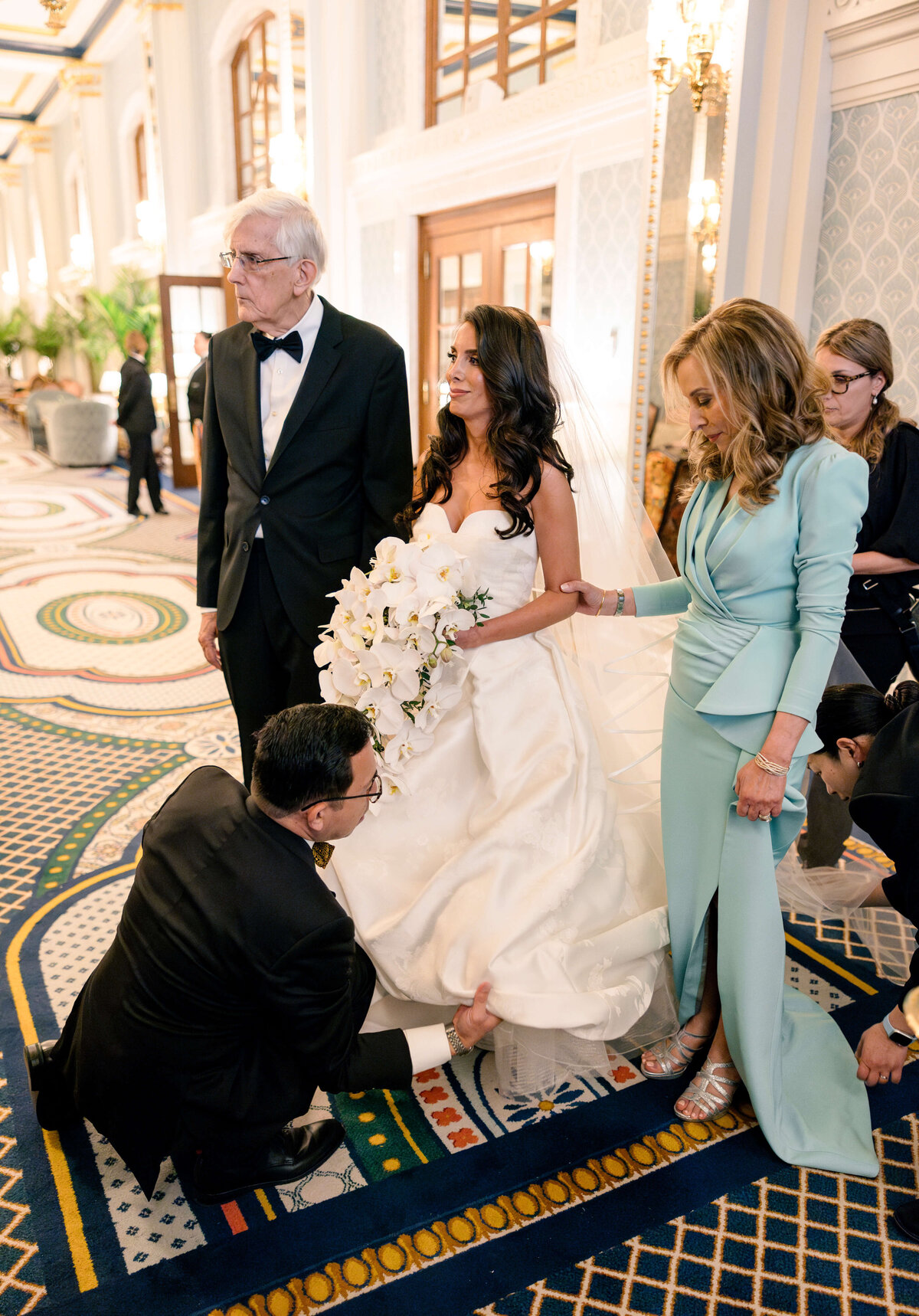 A bride in a white gown holds a bouquet of white flowers, standing beside an older man in a black tuxedo and a woman in a light blue dress. A person kneels, adjusting the brides dress in a lavishly decorated room.