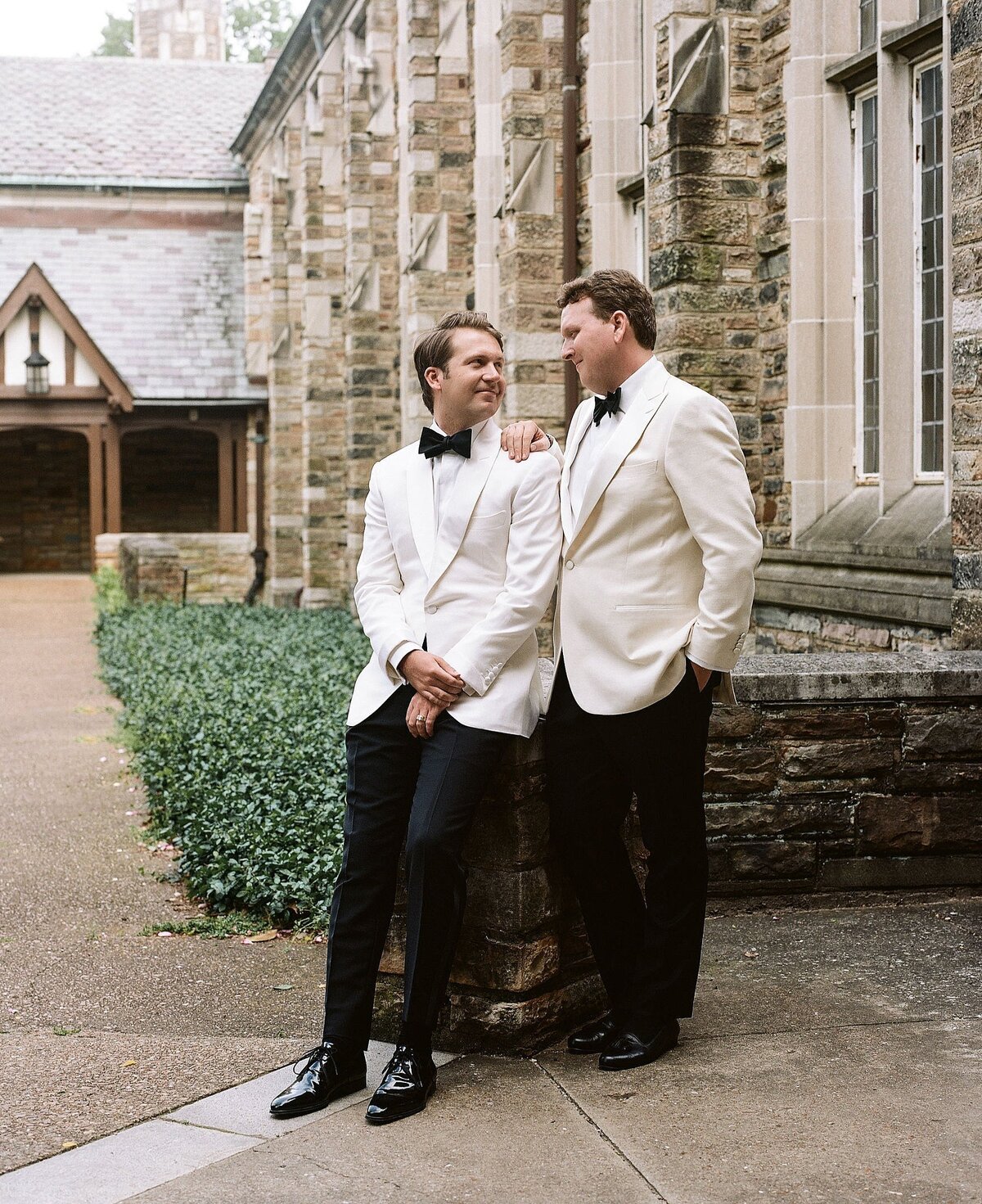 Two grooms wearing white tuxedo jackets and black pants smile at each other outside a stone church at Scarritt Bennett in Nashville, TN