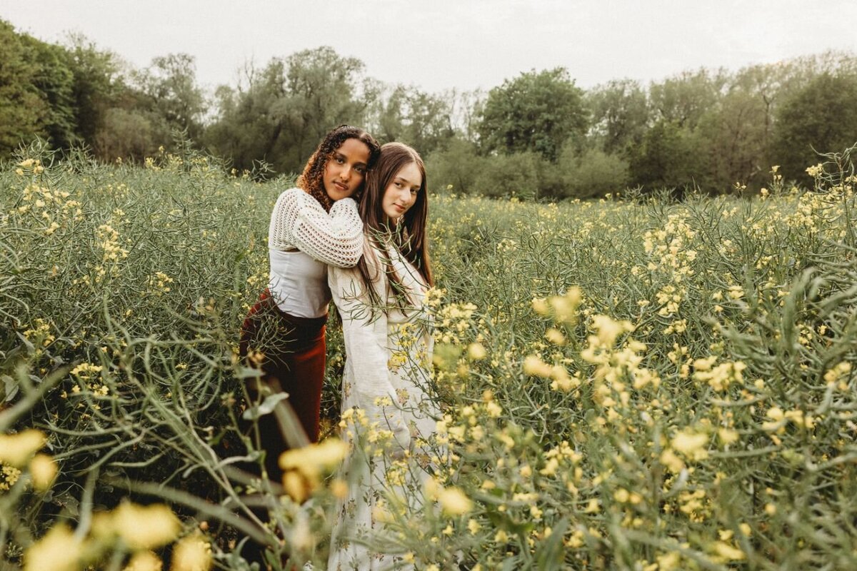 two stylish young women with contrasting hair textures stand affectionately embraced in a field of blooming yellow flowers