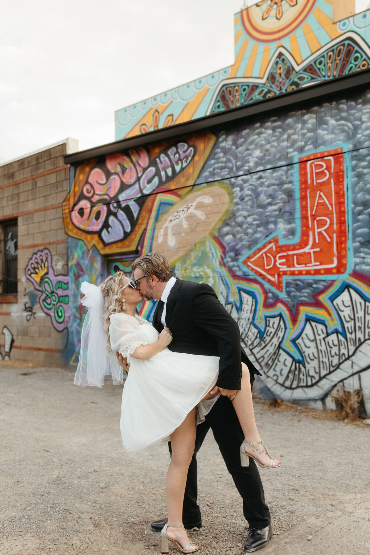 An exuberant couple in wedding attire sharing a kiss in front of a colorful graffiti wall