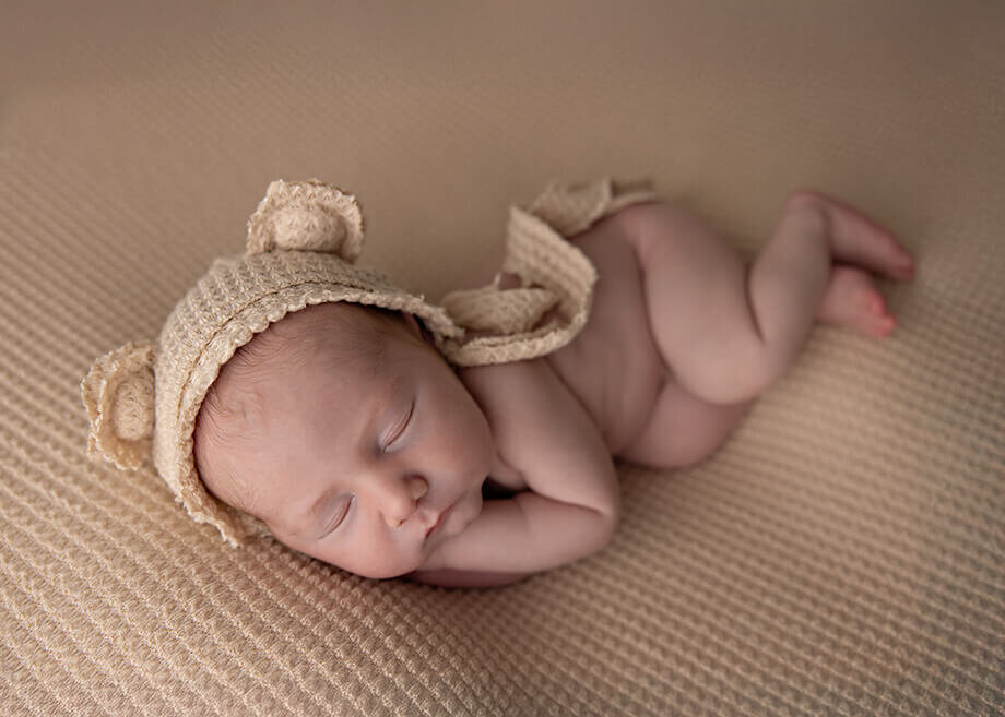 Newborn Boy laying on his side on a dark beige textured backdrop and a bear bonnet.