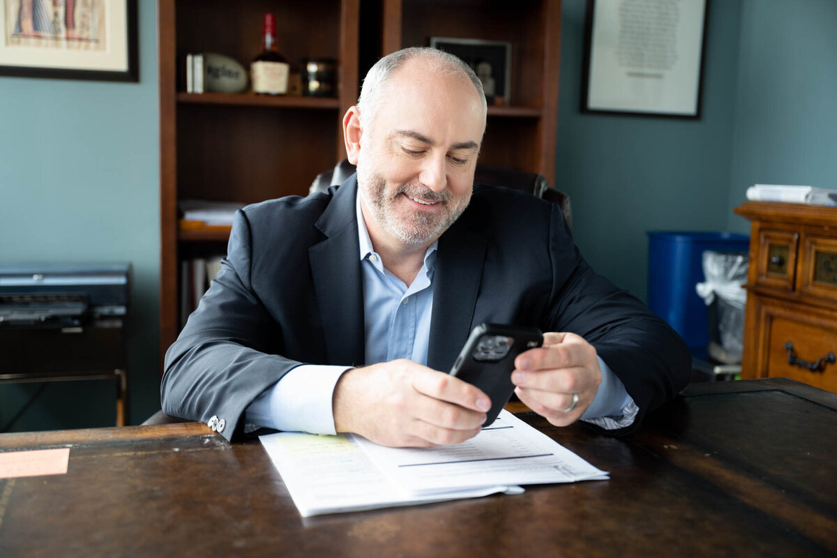 a man sitting behind a desk looking down to his cellphone and smiling
