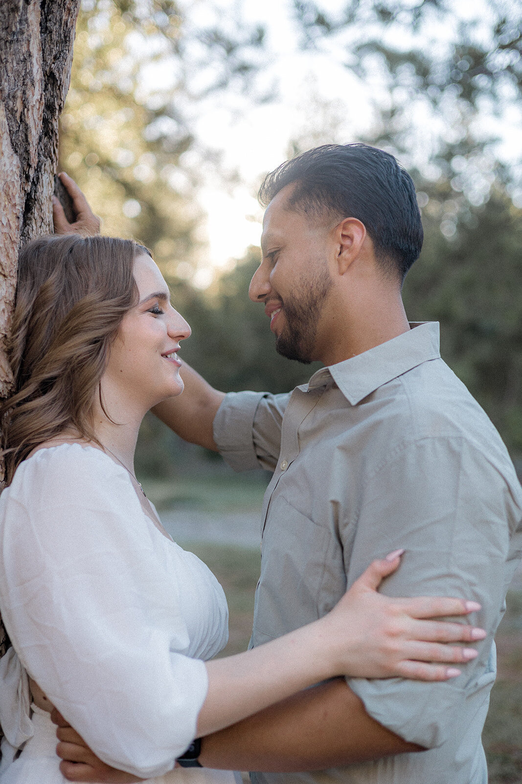 A couple stands closely by a tree, gazing at each other with soft smiles, surrounded by the warm glow of sunlight filtering through nearby branches.