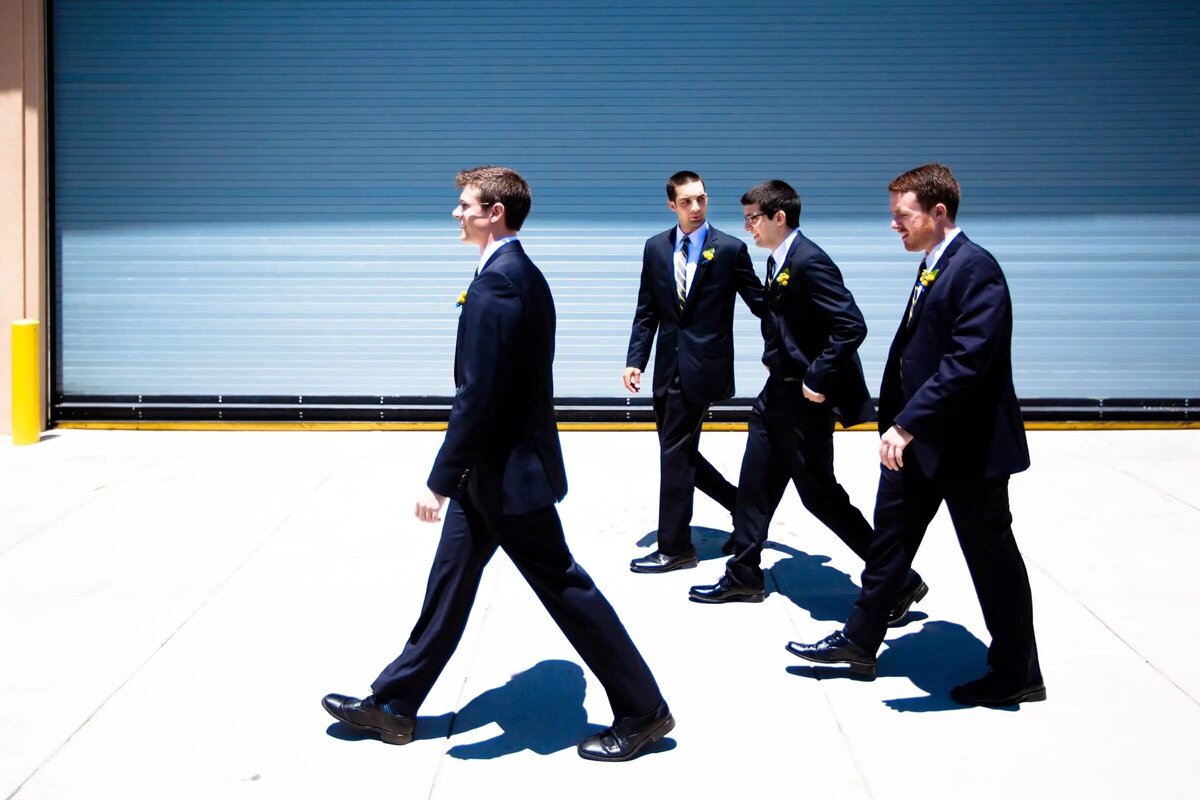 Four groomsmen walk in unison against a bright yellow industrial backdrop, their black suits standing out boldly.