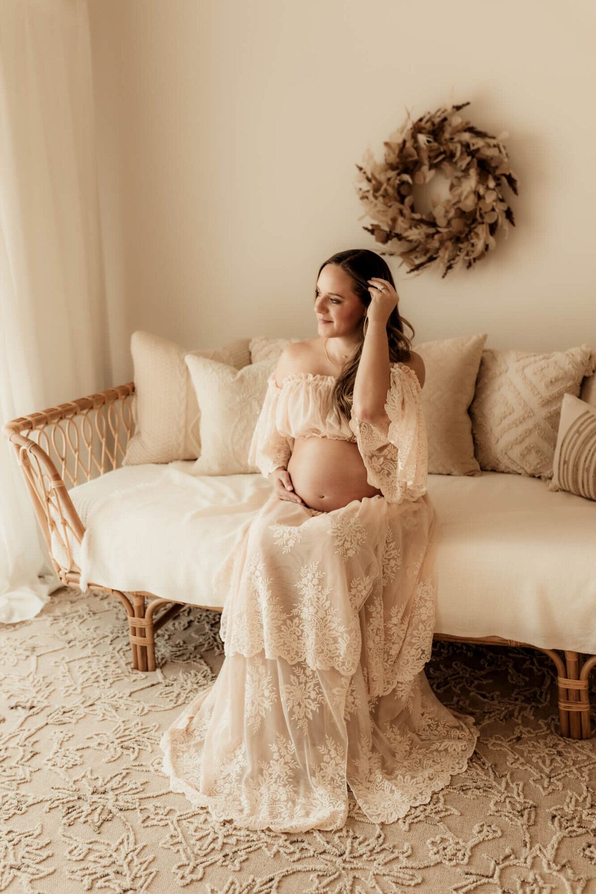 Pregnant mother wearing a blush pink lace dress pulls her hair behind her ear while sitting down for a studio photo in OKC.