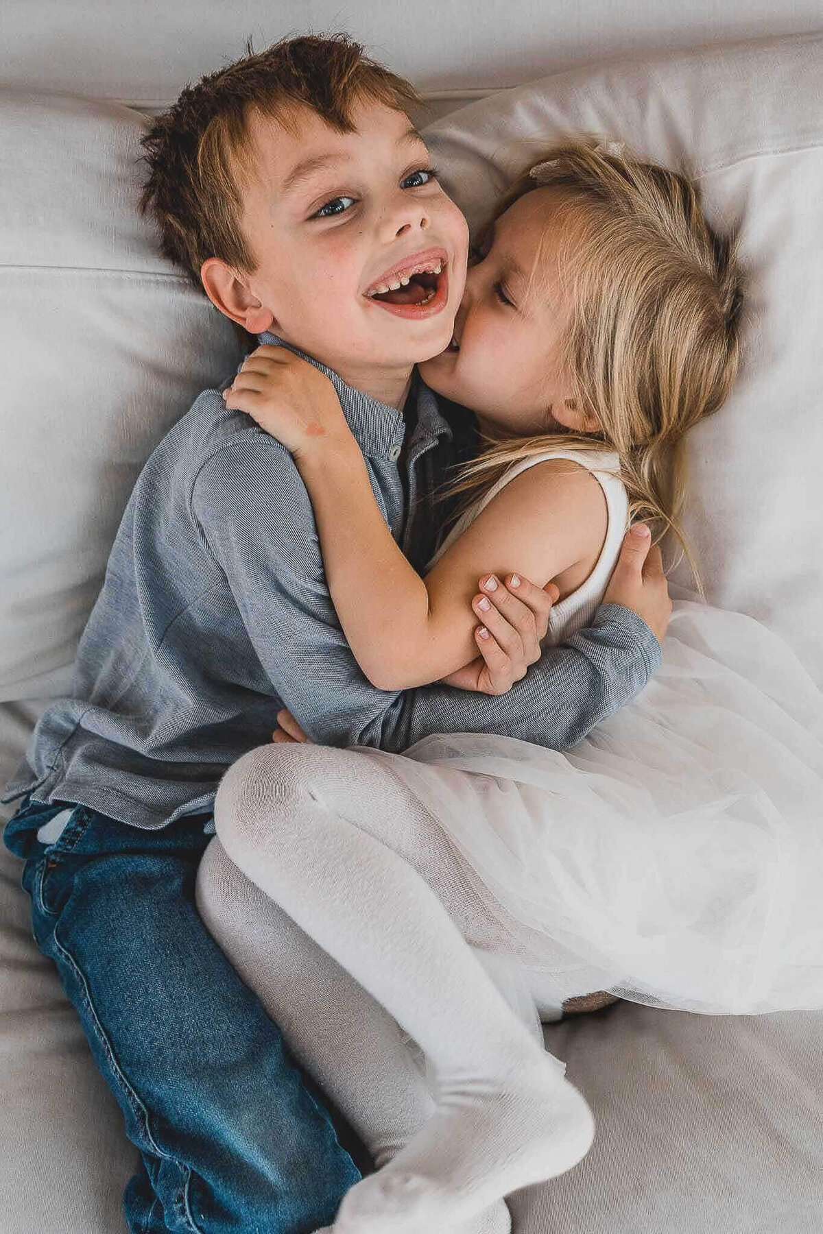 Brother and sister laughing on white couch during in-home family photography session.
