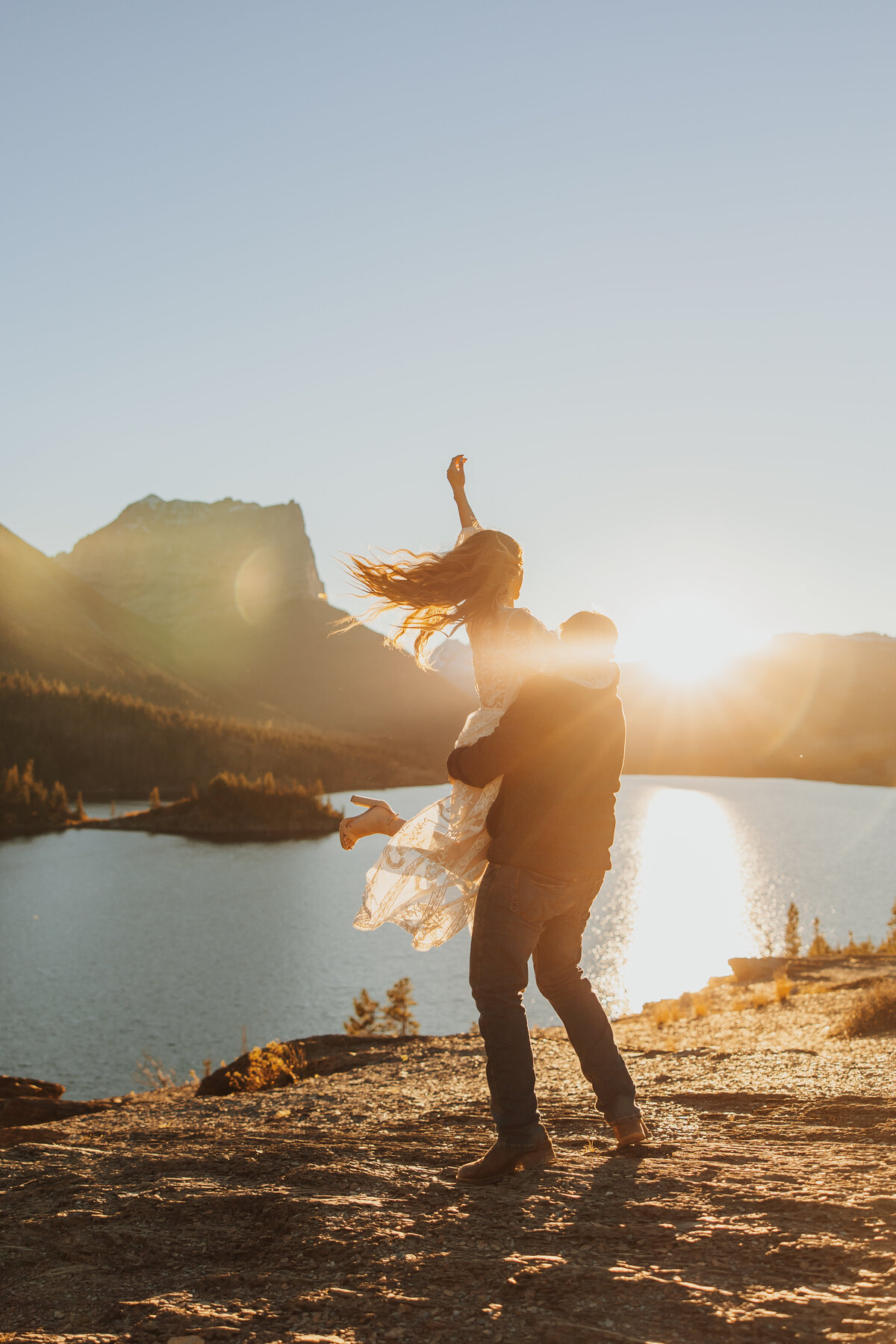 Groom spinning bride around joyfully in Glacier National Park with a lake in the background.