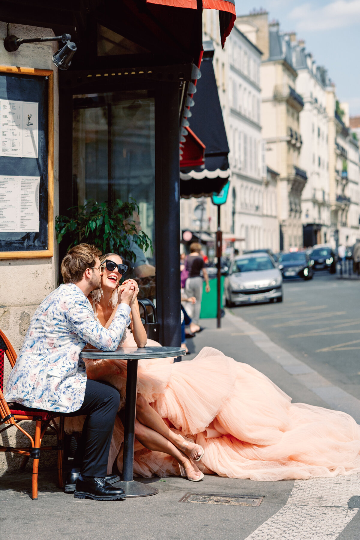 Couple at a cafe enjoying champagne, she in a peach gown with long train at their Paris engagement session, photographed by Italy wedding photographer.