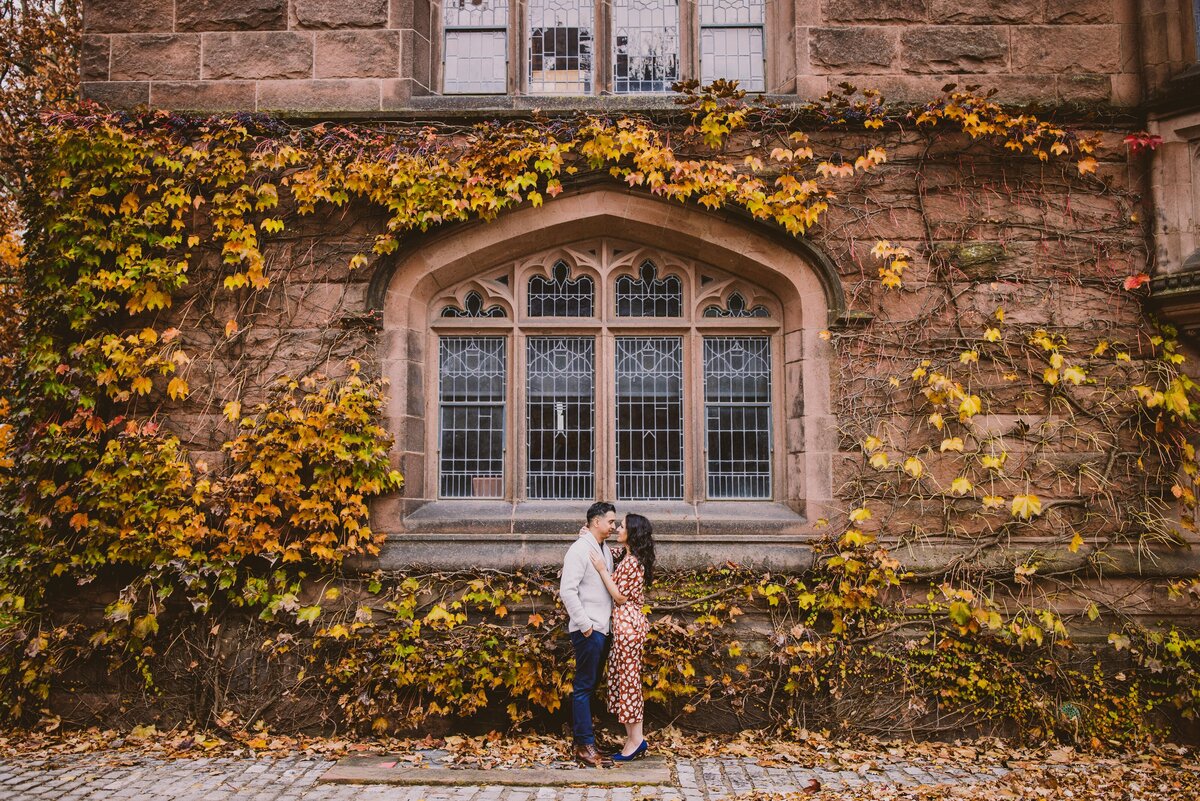 A couple standing in front of a window with fall leaves on the wall at princeton university in nj