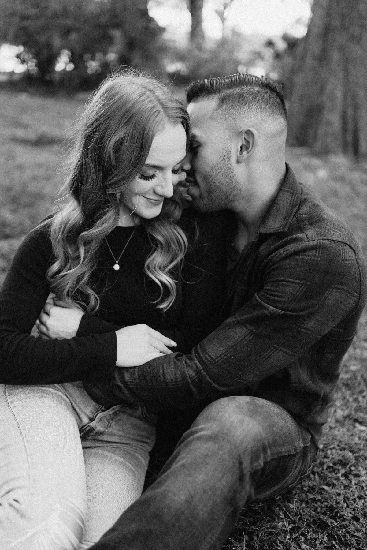 Black and white photo of engaged couple casually leaning into each other on the grass while the man holds the woman and appears to whisper in her ear. Photo taken by Orlando Wedding Photographer Four Loves Photo and Film.