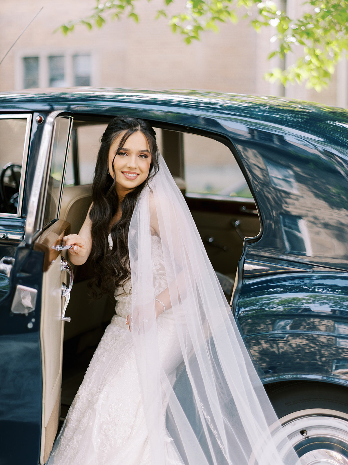 A bride in a white dress and veil is stepping out of a classic black car, holding the door handle. Trees and a building are visible in the background, capturing the essence of an elegant wedding at Fairmont Palliser Calgary.