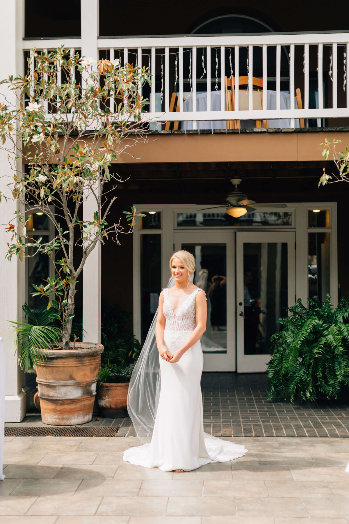 A bride in a white dress and veil stands smiling outdoors in front of double doors, beneath a balcony, beside large potted plants, with sunlight illuminating the scene.